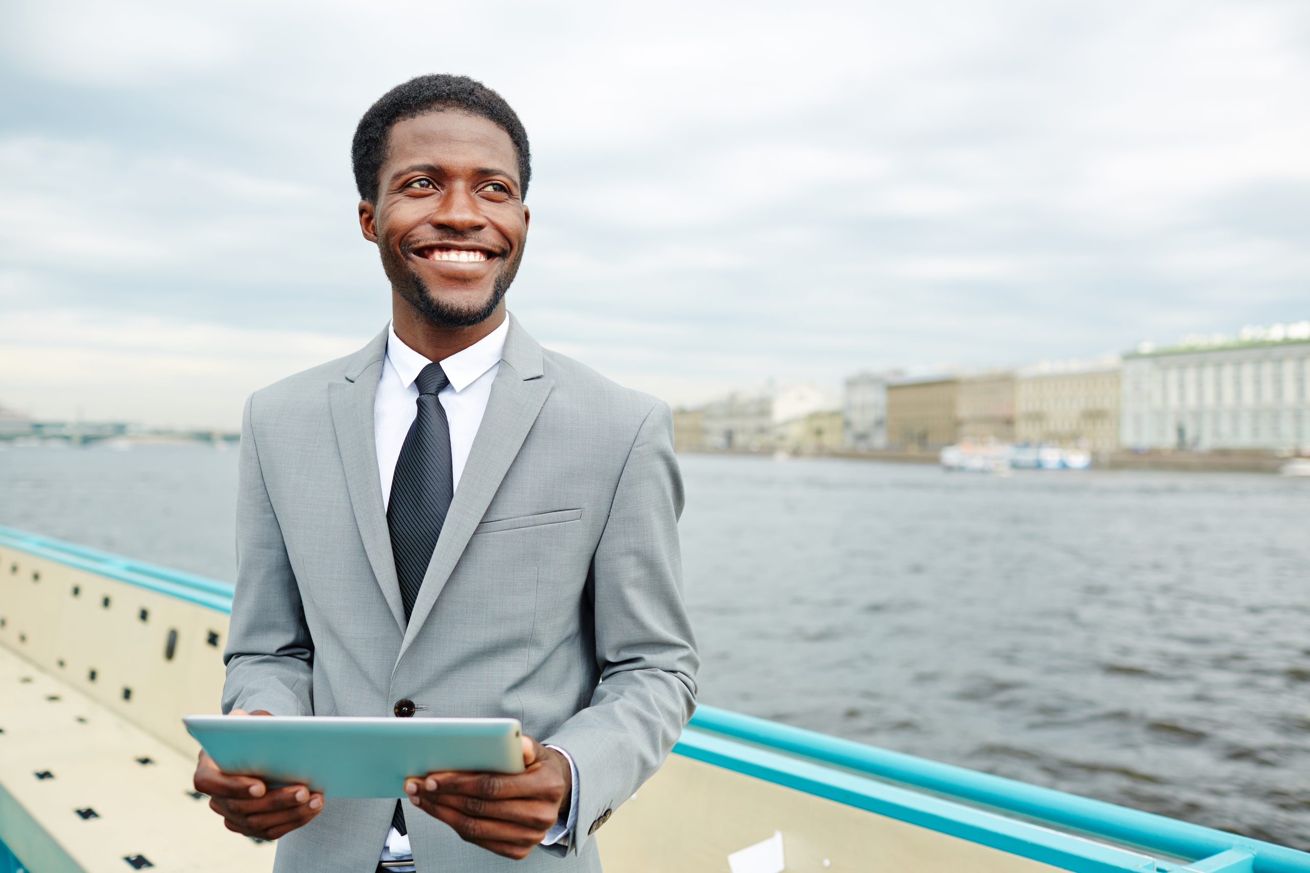 Handsome African American manager looking away with toothy smile while standing on upper deck of ship and checking business emails with help of digital tablet, waist-up portrait shot